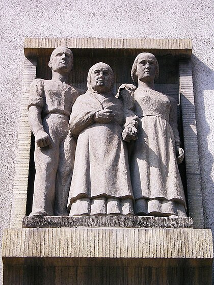 Steinrelief von 1938 über dem Eingang: ein junger Mann, eine alte Frau, eine junge Frau