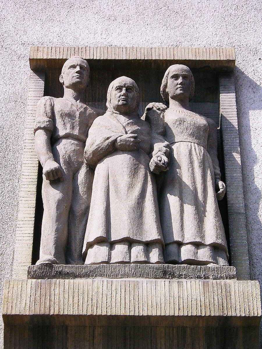 Das Steinrelief über dem Eingang: links ein junger Mann in Arbeitskleidung, in der Mitte eine alte Frau mit Kopftuch, rechts eine junge Frau, die sie hält