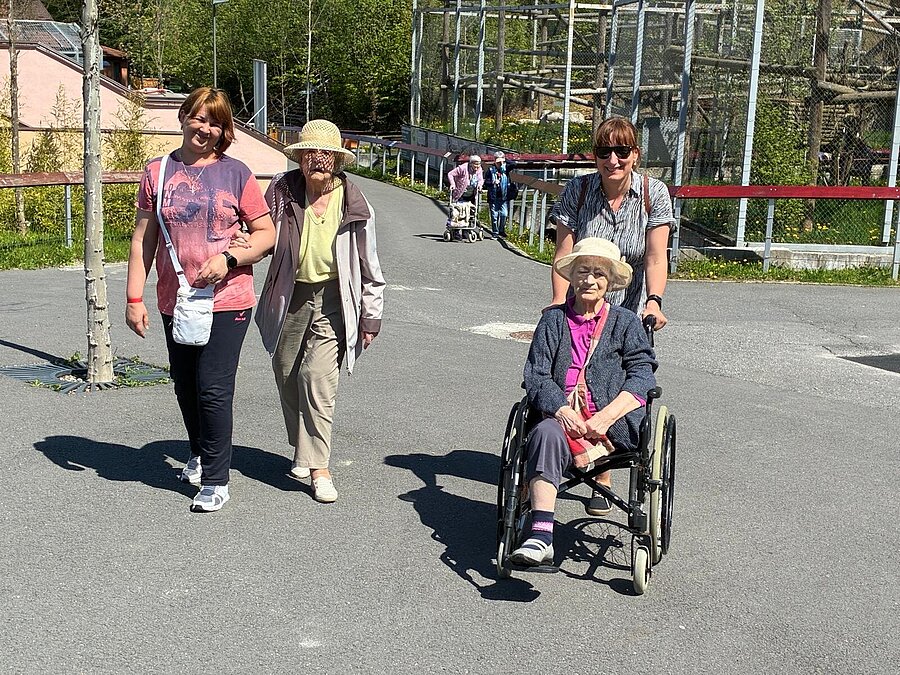 Zwei Bewohnerinnen zu Fuß und im Rollstuhl mit Mitarbeiterinnen auf dem Weg durch den Zoo
