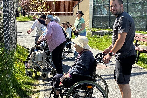 Bewohner im Rollstuhl vor einem Gehege, ein Mitarbeiter schiebt